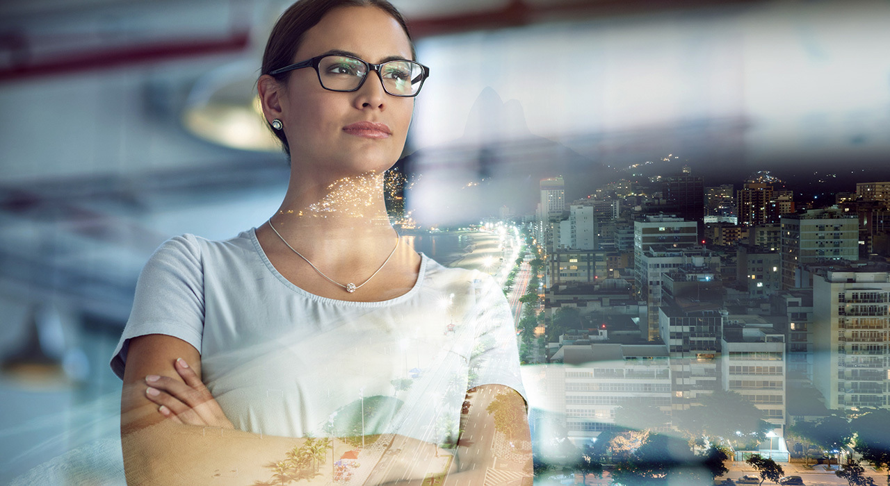 A woman in glasses with her arms crossed holds a determined look while starring out at the city from her office.
