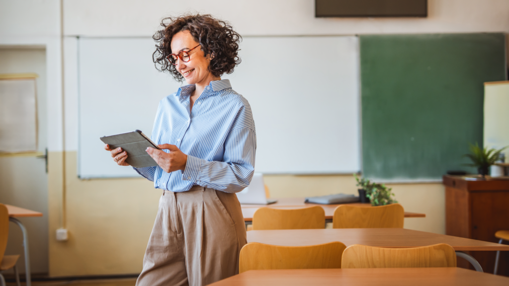 Female teacher with curly hair and glasses holding tablet in empty classroom with whiteboards and student desks