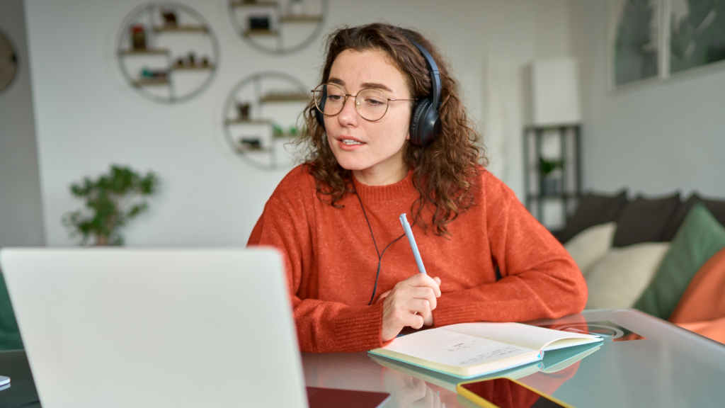 Young woman with headphones and glasses taking notes during online class on laptop at home