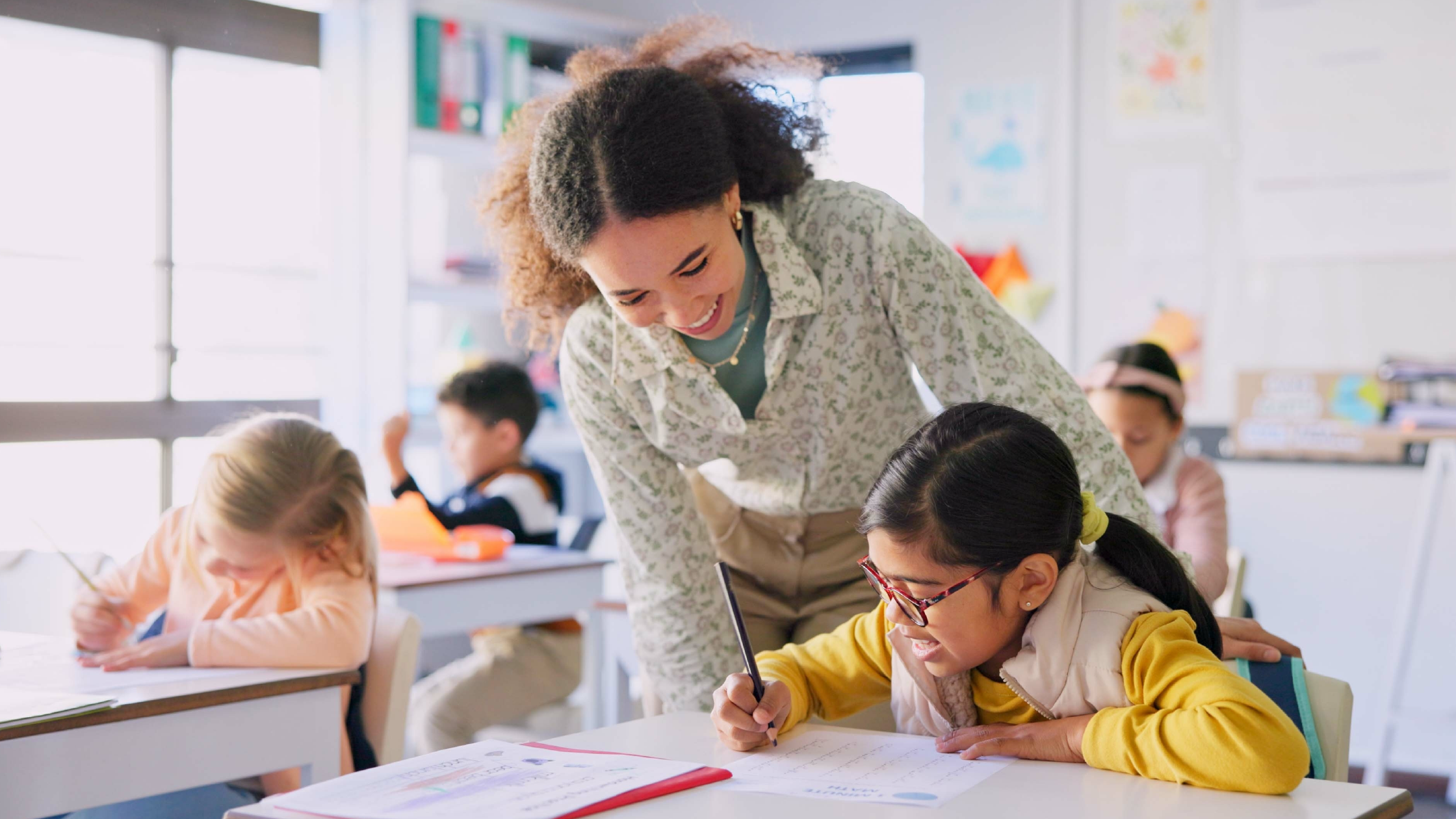 Smiling K-12 teacher leaning over to help young girl with glasses write at her desk in a bright elementary classroom
