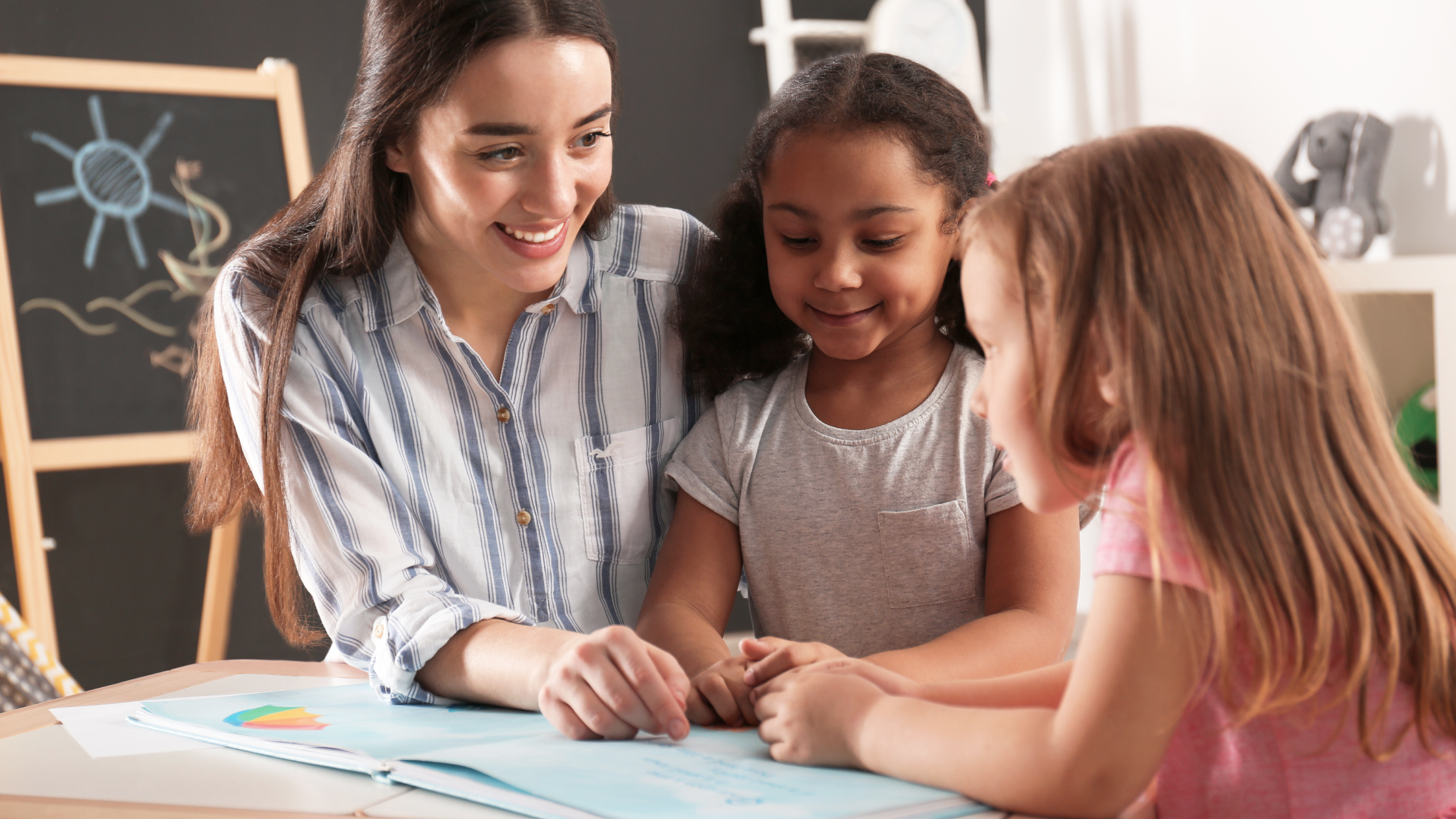 Smiling female teacher reading a colorful book with two young girls at a classroom table
