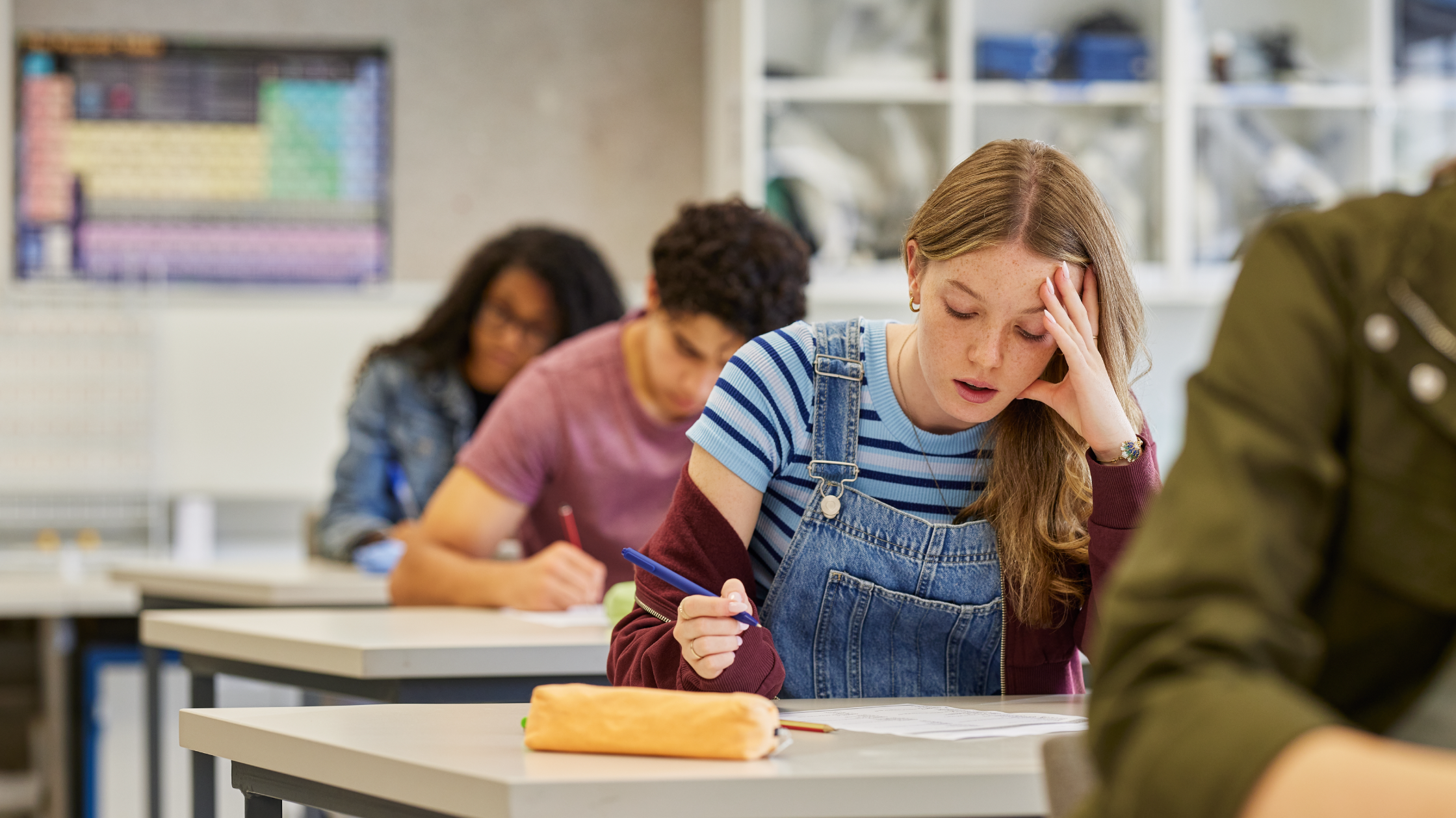 Exhausted teenage girl resting head on hand at school desk during class, struggling to focus
